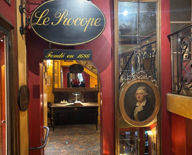 “Interior kafe Le Procope di Paris dengan meja marmer, lampu kristal, dan potret tokoh sejarah di dinding.”