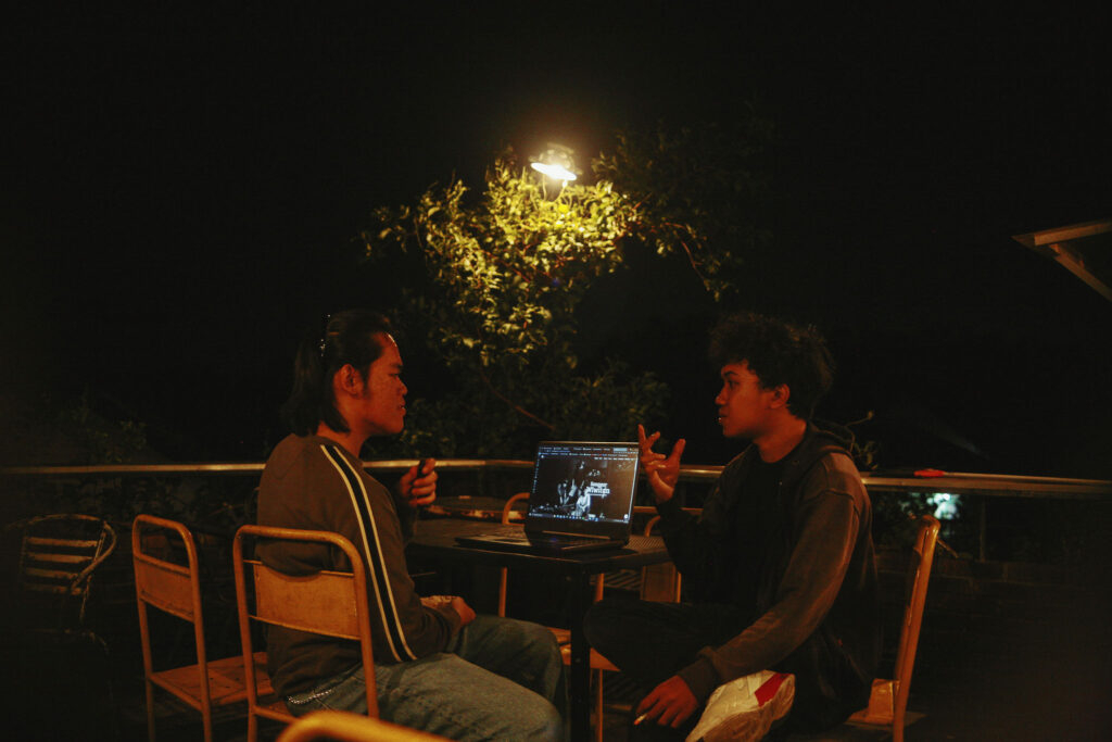 title="Esai senja tentang kopi dari kebun warga, ingatan sejarah, dan bisikan kekuasaan yang jarang dibicarakan." Dua anak muda duduk berhadapan di meja teras pada malam hari, berbincang serius di bawah lampu taman yang temaram Dongeng Kopi