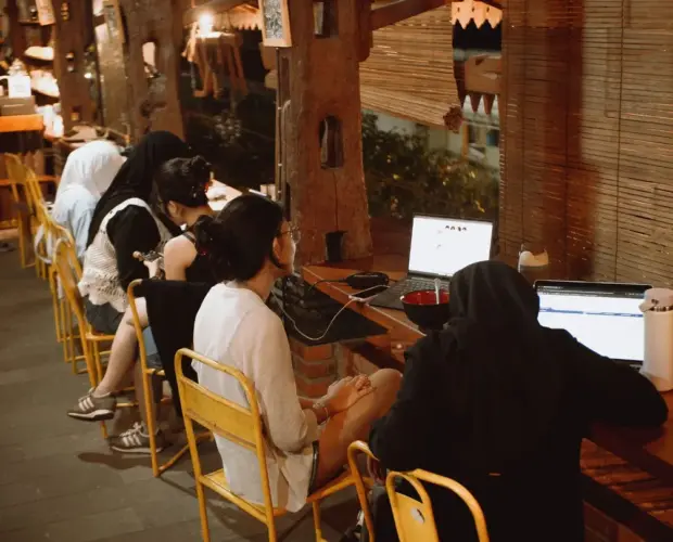 oto suasana kedai dari sudut agak jauh (wide shot) yang memperlihatkan jajaran orang sedang fokus membuka laptop dengan latar belakang rak buku panjang yang temaram dan hangat.